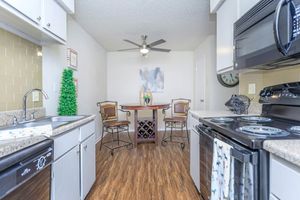 A bright and modern kitchen featuring granite countertops, white cabinets, and stainless steel appliances. There is a small dining area with a round table and two chairs, a wall-mounted clock, and decorative elements like a small green plant and a wall art piece. The flooring is a warm wooden texture.