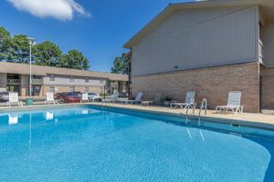 A clear blue swimming pool surrounded by lounge chairs, with a brick building in the background. The scene includes trees and a blue sky with a few clouds, creating a relaxed, inviting atmosphere. Cars are parked nearby, indicating a residential or apartment setting.