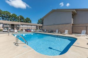 A clear blue swimming pool surrounded by lounge chairs, with a light-colored building in the background. The sky is bright with a few white clouds, and trees are visible beyond the pool area. The pool has a ladder for entry, and the area is well-maintained.