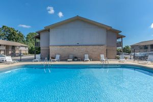 A clear blue swimming pool in the foreground with lounge chairs around it, set against a backdrop of residential buildings under a bright blue sky. The area is well-lit and inviting, with a calm atmosphere perfect for relaxation or enjoying a sunny day.