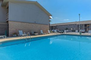A clear swimming pool with lounge chairs around it, set against a brick building and a blue sky. Residential buildings are visible in the background, creating a relaxed outdoor atmosphere.