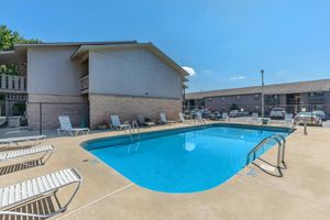 A clear blue swimming pool surrounded by lounge chairs, with a two-story building in the background. The area is sunny with a few cars parked nearby, and a fence enclosing the pool area.