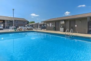 A clear blue swimming pool with a ladder in the foreground, surrounded by a concrete deck. Behind the pool, there are several two-story buildings, some with air conditioning units. The sky is bright with a few fluffy clouds, and the area is fenced. Cars are parked nearby.