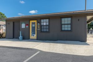 A single-story building with a brown exterior and a bright yellow front door. The facade features windows with black frames, a small concrete area in front, and a blue trash can. A sign is visible next to the door, indicating it serves a specific purpose or business, under a clear blue sky.