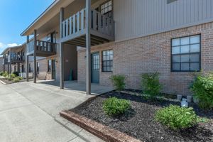 Exterior view of a multistory apartment building featuring a beige brick facade, black-framed windows, and wooden balconies. The pathway is lined with landscaping that includes shrubs and small plants. Clear blue skies above provide a sunny ambiance.