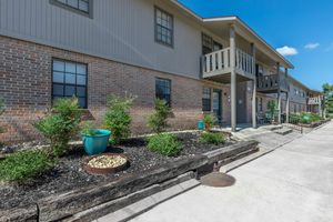 A view of a multi-unit residential building featuring a brick facade, green landscaping, and a concrete walkway. The building has balconies on the upper level, and potted plants are visible near the entrance. The sky is clear and sunny, adding to the inviting atmosphere of the property.