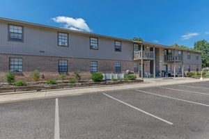 A two-story apartment building with a brick facade and gray siding. The building features multiple windows, a balcony on the upper floor, and a small outdoor seating area. Surrounding landscaping includes shrubs and small plants. Clear blue sky above with a few clouds. Parking spaces are visible in front.
