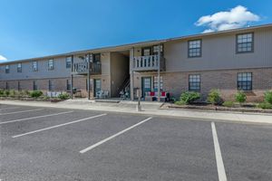 Two-story apartment building with a brick facade and gray siding. It features balconies with white railings. In front, there is a parking lot with several empty spaces, and landscaped areas with shrubs. The sky above is blue with scattered clouds.