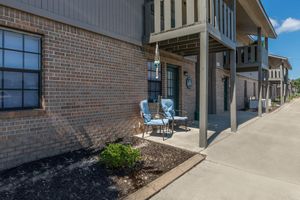 A residential building features a sidewalk in front, with two blue chairs and a small table set up on a covered porch. Decorative elements include a hanging wind chime and a wall clock. The exterior is made of red brick, with windows framed in black. Lush mulch and small landscaping are visible in the foreground.