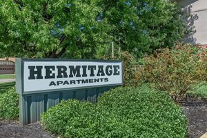 Sign for Hermitage Apartments with green landscaping and a bush with blooming flowers nearby, set against a bright, sunny background.