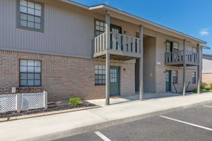 Exterior view of a two-story residential building featuring a brick facade, beige siding, and green doors. Each unit has a small balcony and the entrance is accessed via a set of steps. A parking lot is visible in front, with a landscaped area including plants and a white fence.