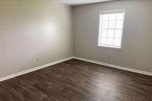 A vacant room with light gray walls and a single window featuring white blinds. The floor is dark wood laminate. There are no furnishings or decorations visible, giving the space a clean and empty appearance.