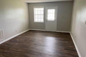 An empty room with light gray walls and a dark wooden laminate floor. There is a window with white blinds on the left, and a door with a small window on the right. The space is spacious and well-lit, giving a feeling of openness and simplicity.