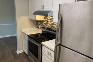 A modern kitchen featuring white cabinets, a stainless steel refrigerator, and an oven with a smooth top. The countertop has a light-colored marble finish, and the backsplash consists of dark and light tiles. The kitchen is well-lit and showcases a clean, contemporary design.