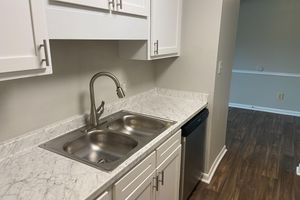 A modern kitchen featuring a double sink with a sleek faucet, white cabinets, and a marble countertop. The view includes a dishwasher and a glimpse of a hallway leading to another room, with light-colored walls and dark wooden flooring.