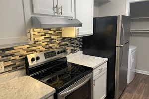 A modern kitchen featuring white cabinets, a stainless steel stove with an overhead range hood, and a black refrigerator. The backsplash consists of decorative tiles, and the countertops are made of light-colored marble. A laundry area with a washer and dryer is visible through an open door in the background.
