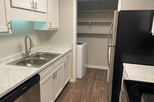 A clean and modern kitchen featuring white cabinets, a double sink with a chrome faucet, and a marble countertop. To the right, there is a black refrigerator and a stove with an oven. A laundry area is visible in a closet space at the end of the kitchen, equipped with a washing machine and open shelving.
