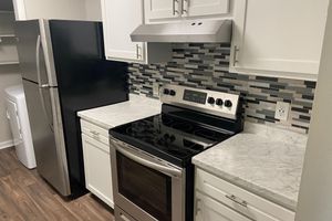A modern kitchen featuring white cabinets, a black refrigerator, a silver range hood above an electric stove, and a marble countertop. The backsplash consists of gray and black tiles, and the floor is made of dark wood. A laundry area is visible in the background with a washer and dryer.