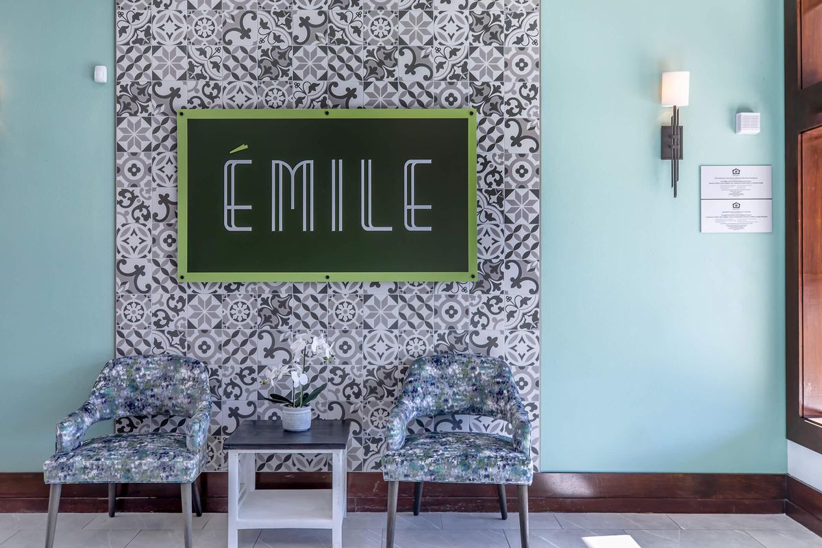 A stylish reception area featuring a patterned tiled wall with a green sign that reads "ÉMILE." There are two patterned chairs on either side of a small table with a potted plant. Soft lighting from wall sconces enhances the inviting atmosphere.