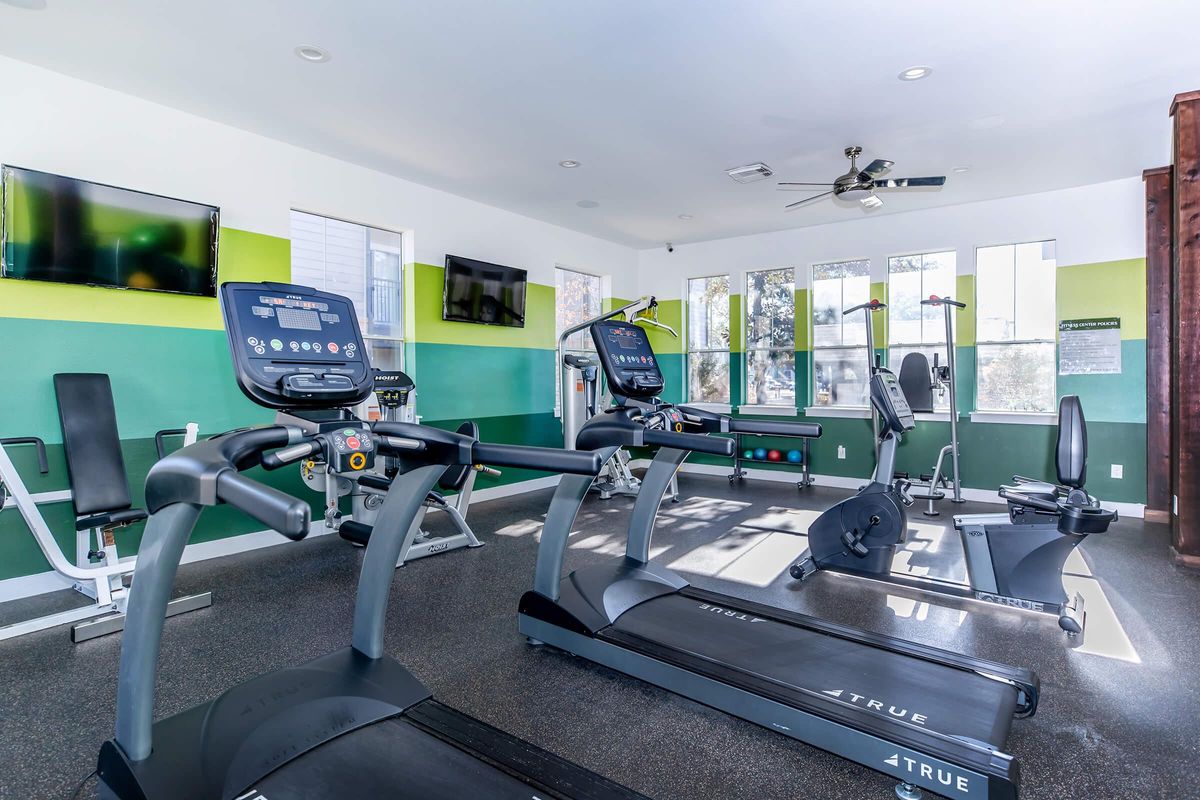 A bright and modern gym space featuring several fitness machines, including treadmills and stationary bikes. The walls are painted in green and white, and large windows provide natural light. TV screens are mounted on the walls for entertainment. The flooring is dark and suitable for workouts.