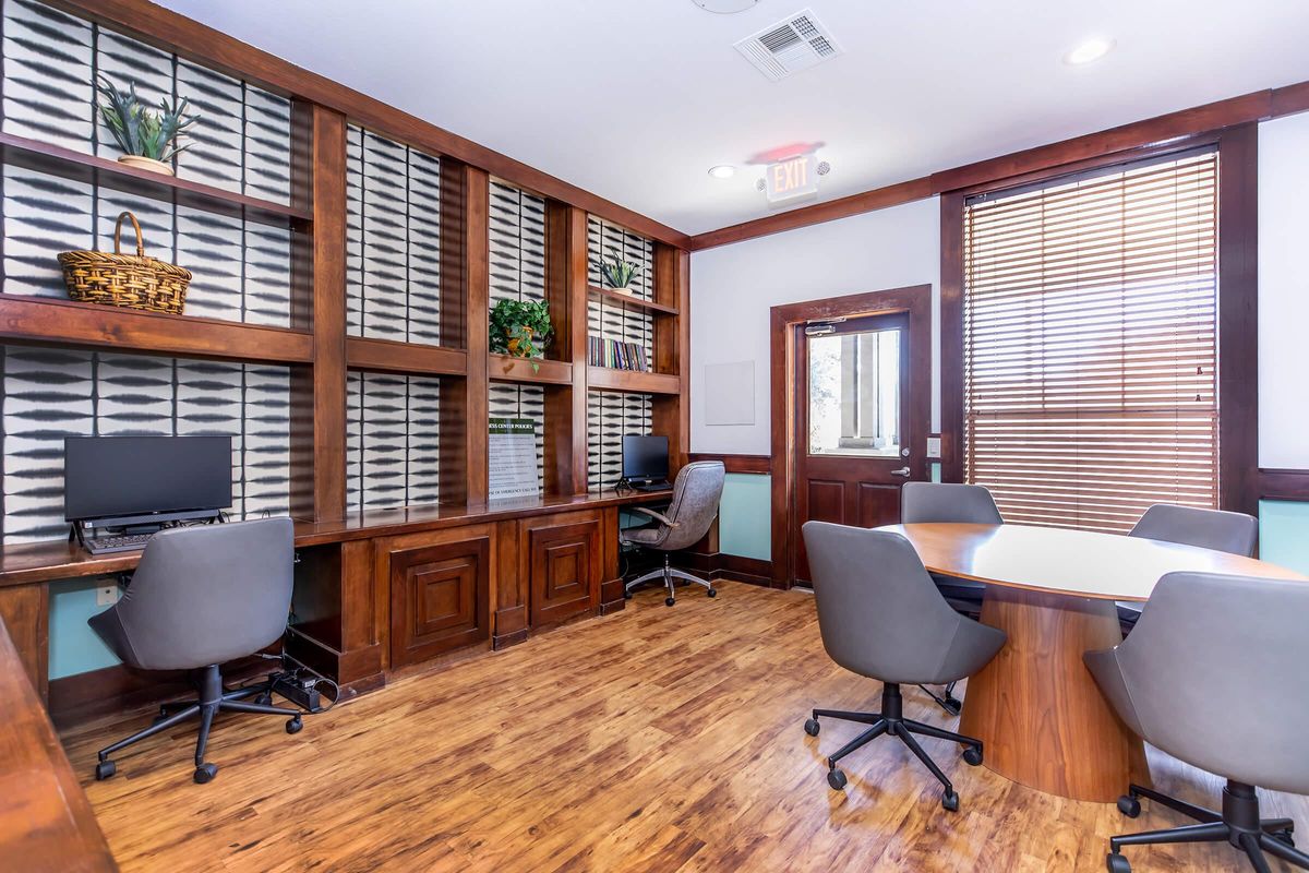 A modern office space featuring wooden accents, a large table surrounded by gray office chairs, and two computer workstations in built-in shelves. Natural light filters through blinds, providing a bright and inviting atmosphere suitable for meetings or collaborative work.