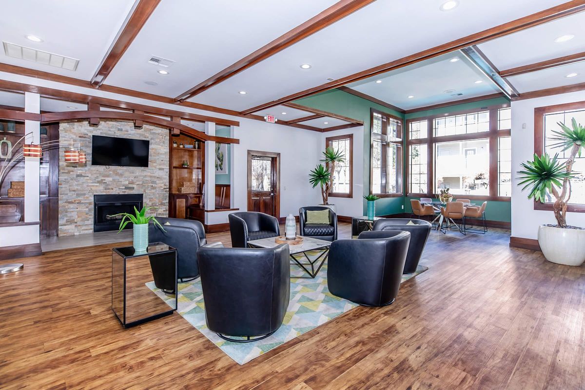 A modern lounge area featuring four black armchairs arranged around a glass coffee table. A decorative rug lies beneath them, and there are large windows allowing natural light. In the background, there is a stone fireplace, and potted plants are placed around the space, giving a welcoming atmosphere.