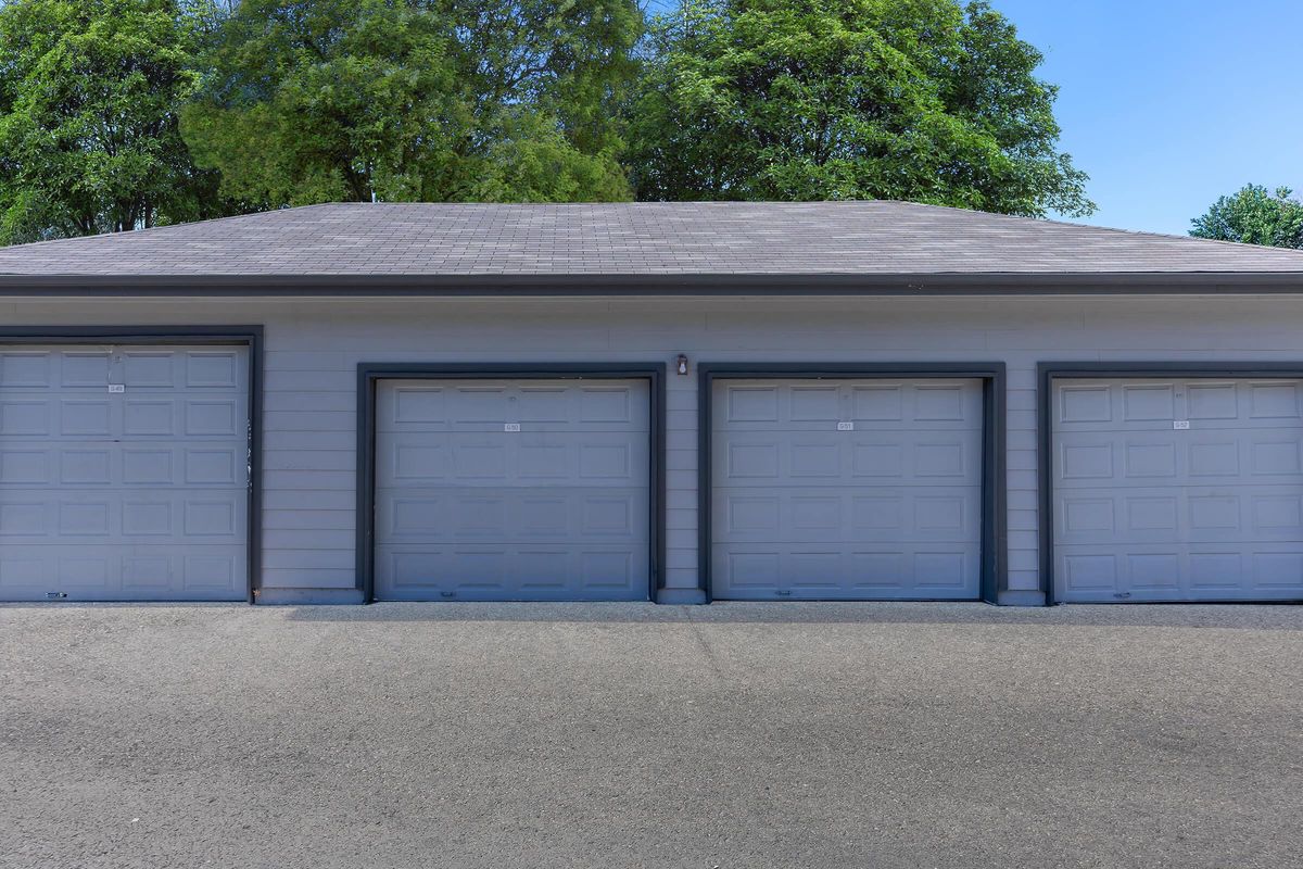 Three gray garage doors lined up on a light-colored building, with a smooth driveway in front and green trees in the background. The sky is clear and blue, providing a bright, sunny ambiance.