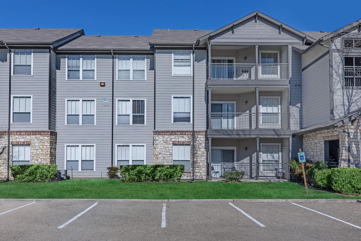 Modern multi-story apartment building with a stone and siding exterior. The facade features multiple balconies, large windows, and a well-maintained lawn. Parking spaces are visible in the foreground, indicating a residential area. Bright blue sky enhances the overall appearance of the property.