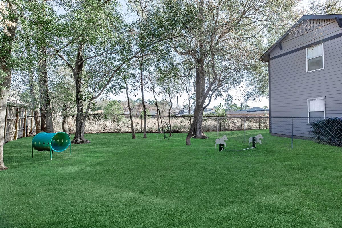 A spacious backyard featuring a well-maintained lawn, a small green tunnel for pets, two playful horse-shaped structures, and a wooden fence. The area is shaded by trees, with a house visible in the background and a clear sky above.