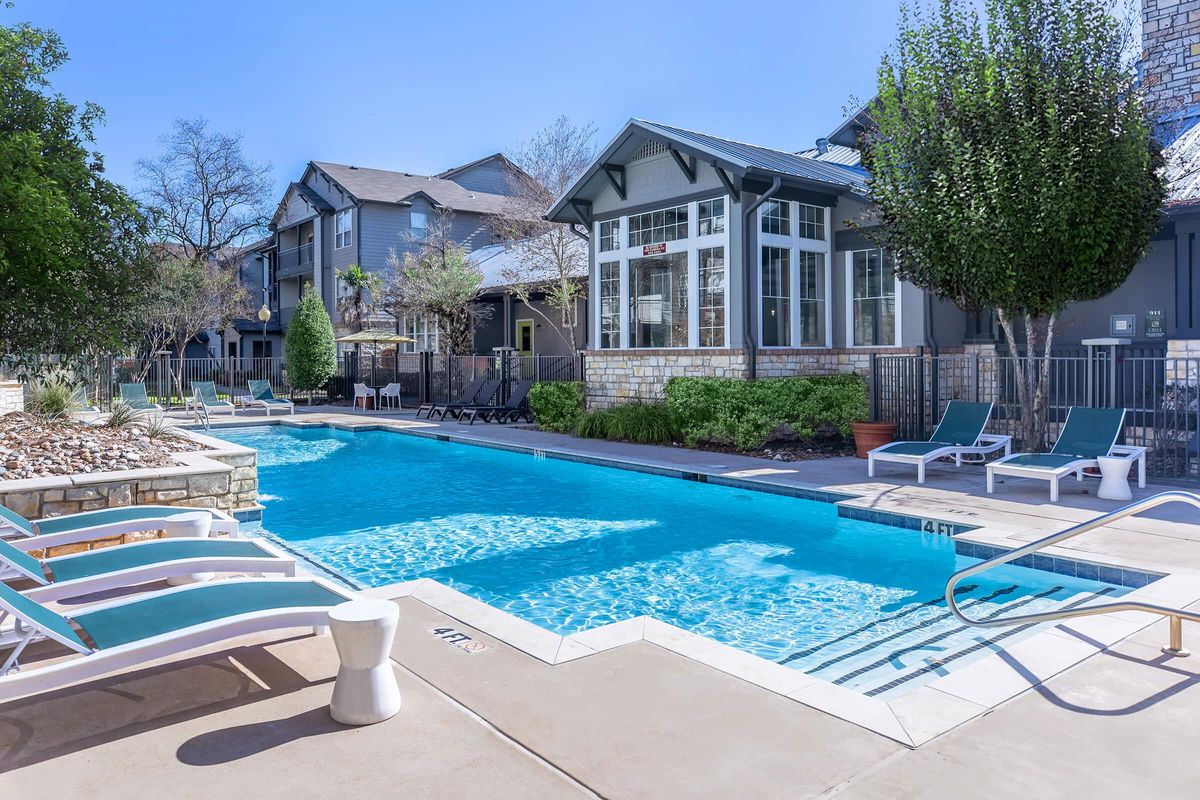 A clear swimming pool surrounded by lounge chairs and potted plants. In the background, there are modern residential buildings with large windows, and the area is filled with greenery under a bright blue sky. The scene is inviting and well-maintained, ideal for relaxation and leisure.