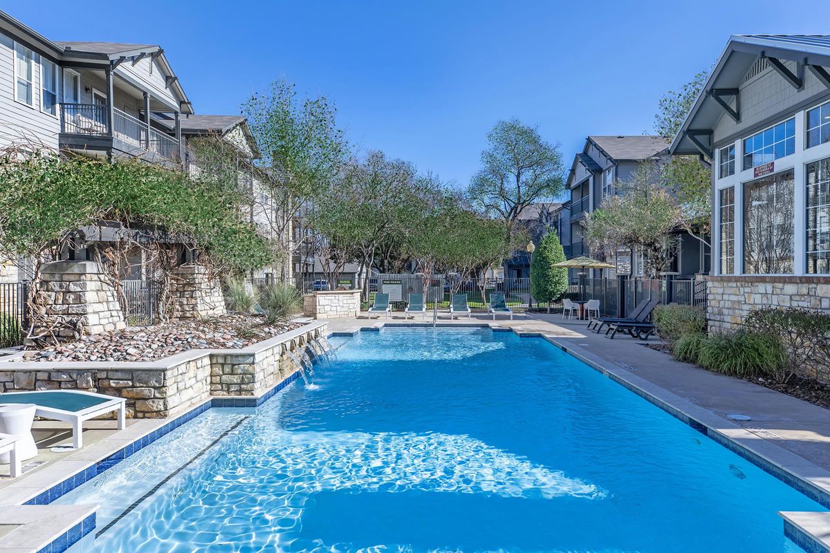 A tranquil apartment community pool area surrounded by landscaped gardens and lounge chairs. The clear blue water of the pool reflects the sunlight, with a serene atmosphere created by the nearby trees and buildings. Ideal for relaxation and leisure.