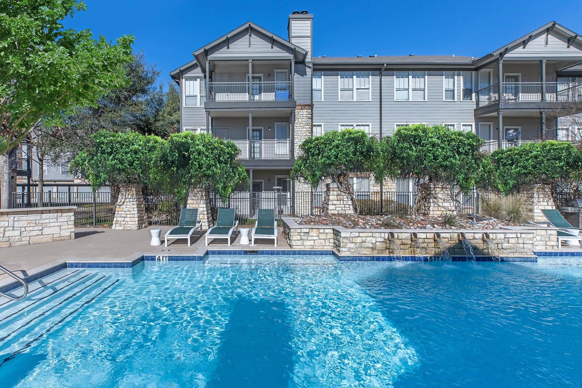 A clear blue swimming pool surrounded by lounge chairs and lush greenery. In the background, a three-story apartment building features several balconies and a well-maintained exterior. The sky is bright blue, indicating a sunny day.