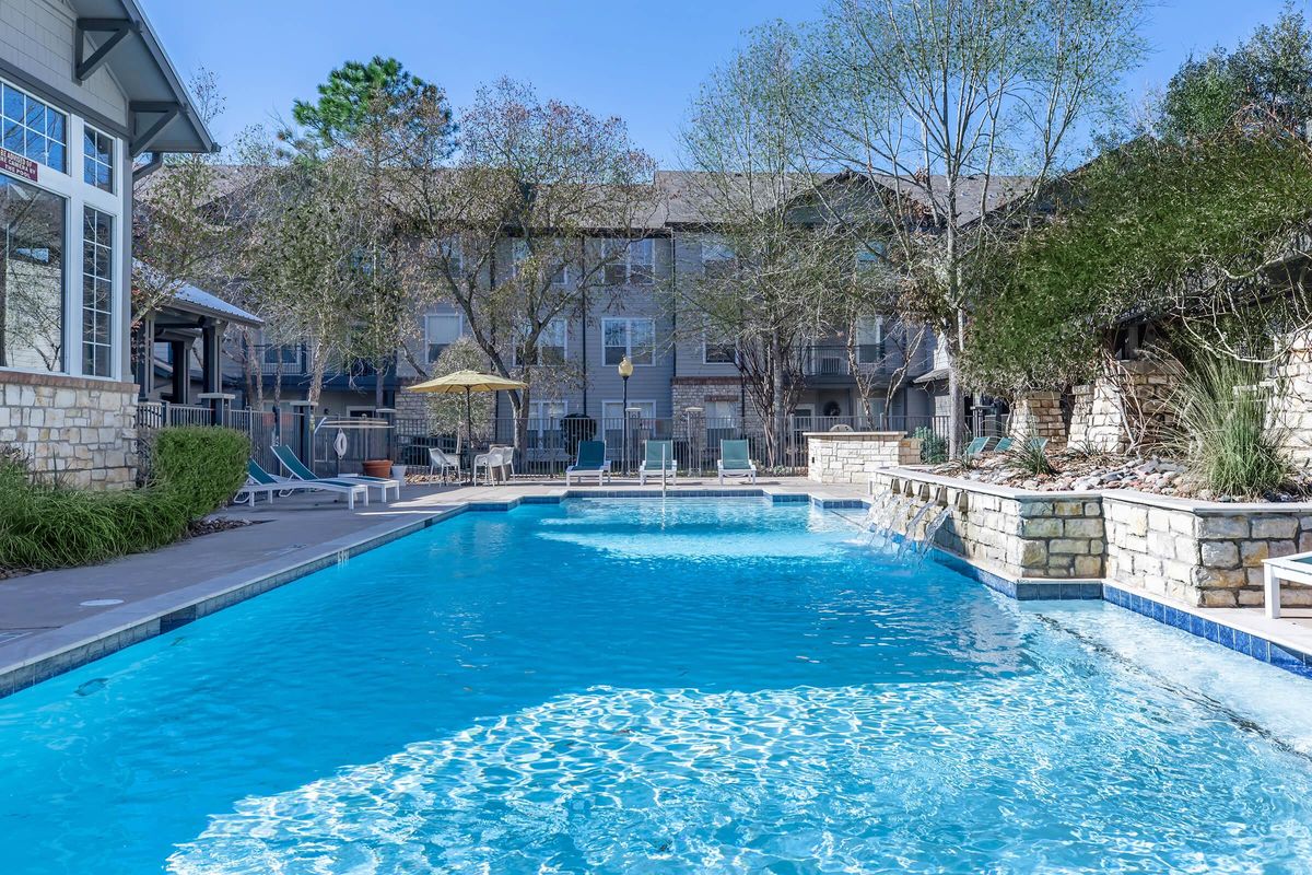 A clear blue swimming pool surrounded by lounge chairs and an umbrella, set amidst landscaped greenery. In the background, there are several multi-story apartment buildings with balconies. The scene is illuminated by bright sunlight, suggesting a warm, inviting atmosphere.