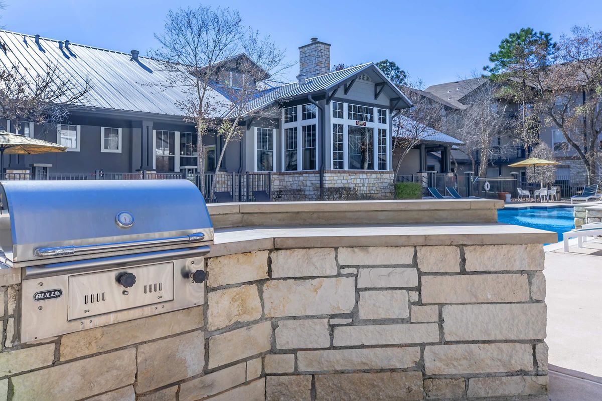 A stone barbecue grill is positioned beside a swimming pool. In the background, there are modern apartment buildings with large windows and a patio area featuring sun umbrellas. The scene is set on a sunny day with clear blue skies and bare trees, indicating early spring or late winter.