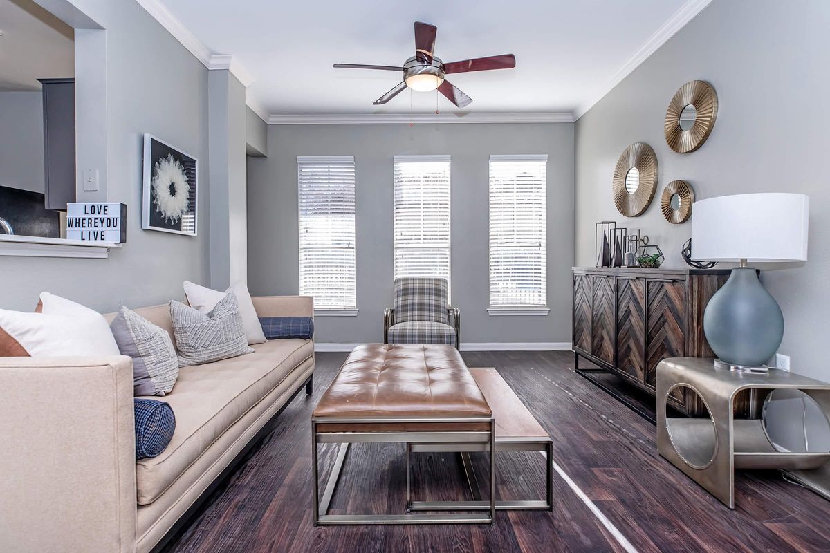 A stylish living room featuring a beige sofa with decorative pillows, a brown tufted coffee table, a light fixture with a fan, and a decorative side table. Large windows with blinds allow natural light, and the walls are adorned with round mirrors. A patterned armchair adds character to the space.