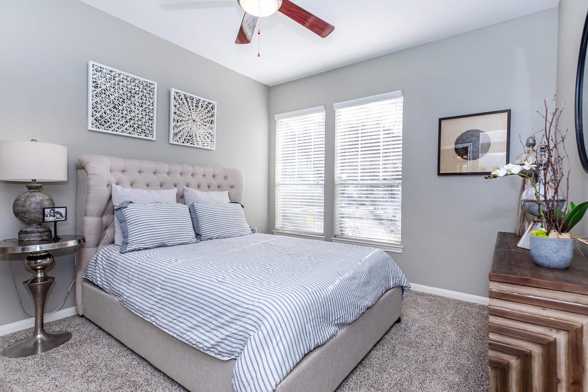 A well-decorated bedroom featuring a beige upholstered bed with striped bedding, flanked by two large windows with white blinds. There's a stylish side table with a lamp and decorative items, along with framed artwork on the walls, and a wooden dresser on one side, enhancing the room's cozy and modern atmosphere.