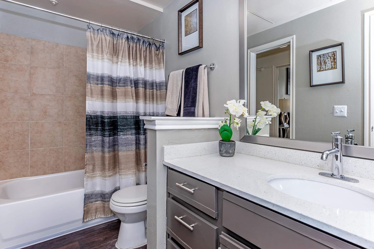 A clean bathroom featuring a beige tiled shower area with a striped curtain, a white bathtub, a modern sink with a marble countertop, and dark cabinetry. Decor includes a small potted plant and framed artwork on the walls, along with towels neatly arranged.