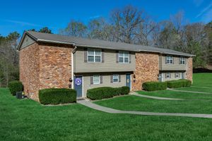 A two-story residential building with a combination of brick and siding exterior. The building features multiple units, each with a door and framed by neatly trimmed bushes. The well-maintained green lawn surrounds the property, and tall trees are visible in the background under a clear blue sky.