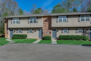 A two-story tan apartment building with red brick accents. There are four doors on the front, two on each level, surrounded by neatly trimmed green shrubs. The building is set against a clear blue sky with trees in the background. A paved pathway leads from the parking area to the entrance.