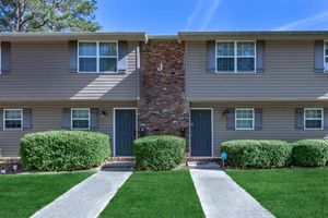 Two identical townhouse units with gray siding and dark green doors, separated by a brick column. Each unit has a small porch and two windows on the upper floor. The front yard features well-manicured green grass and rounded shrubs on either side of a concrete walkway leading to the doors. Clear blue sky above.
