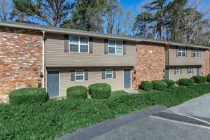 A row of two-story apartment buildings with brick and beige siding. Each unit has a front door with small windows on either side. Lush green shrubs line the walkway, and tall trees are visible in the background under a clear blue sky. The setting appears well-maintained and inviting.