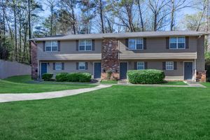 A two-story duplex with tan siding and brick accents, featuring two separate front doors. Neatly trimmed shrubs line the front, and a curved pathway leads to the entrance. Lush green grass surrounds the building and trees are visible in the background under a clear blue sky.