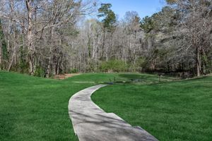 A peaceful landscape featuring a winding concrete pathway leading through lush green grass, surrounded by trees in a forested area. The scene is set under a clear blue sky, showcasing a tranquil outdoor space ideal for walking or relaxing in nature.