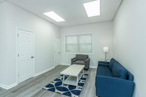 A light and airy room featuring a blue couch, a matching armchair, and a coffee table on a patterned rug. The walls are painted white, and there's a window with blinds allowing natural light in. A floor lamp stands next to the armchair, and there's a closed door visible to the left.