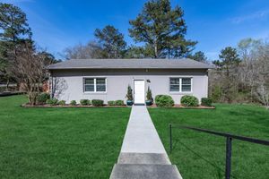 A single-story gray house with a white front door, surrounded by neatly trimmed shrubs and grass. A concrete pathway leads to the entrance, flanked by small planters. The backdrop features tall trees under a clear blue sky.