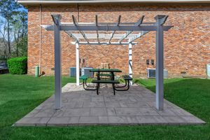 A pergola structure with a picnic table underneath is set on a paved area surrounded by a well-manicured lawn. The backdrop features a brick wall of a building, with greenery on one side and air conditioning units on the other. The scene is illuminated by natural light.