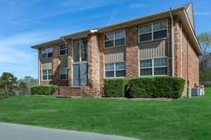 A two-story brick apartment building with large windows, surrounded by neatly trimmed green bushes and grass. The entrance features a set of steps leading to a central door, while the background includes trees and a clear blue sky.