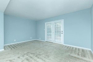 Empty room with light blue walls and beige carpet. There are two sets of glass-paneled double doors leading to a patio or balcony. The room is well-lit with natural light and has a clean, minimalistic look, ideal for decorating or furnishing. No furniture is present.