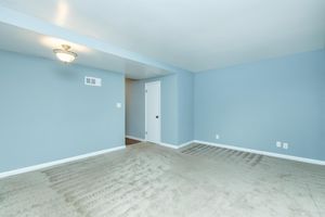 A spacious, empty room with light blue walls and a soft, beige carpet. The ceiling features a decorative light fixture. There is a white door leading to another room, and the overall atmosphere is bright and airy, providing a clean slate for potential furniture and decor.