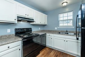 Modern kitchen featuring white cabinetry, a black stove and refrigerator, and stainless steel accents. The countertop is textured in gray, with a double sink positioned under a window that provides natural light. The room has a clean, organized layout with wooden flooring.